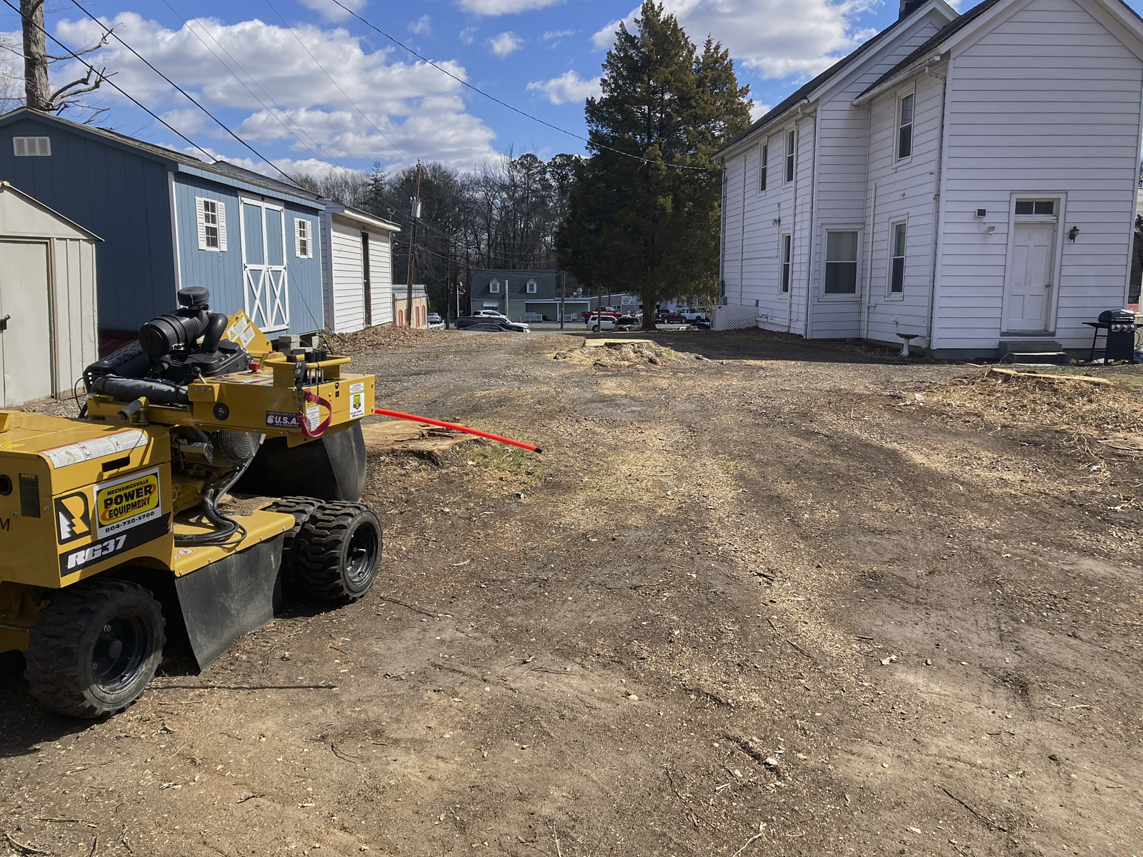 A Long Shot Of A Cleared Ground Area With Some Houses Around Where A Skid Steer Vehicle Is Ready For Tree Stump Removal